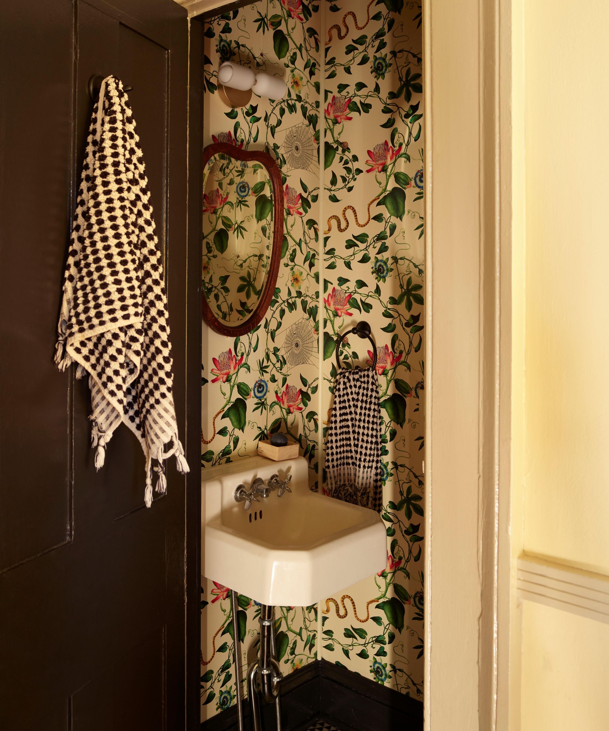 Small powder room with floral and serpent wallpaper, a white pedestal sink, and a black and white patterned towel hanging on a dark brown door