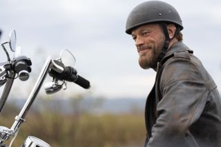 Ares (Adam Copeland) sits on a motorbike (which is out of shot apart from the handlebars) wearing a helmet and a leather jacket and grinning cockily