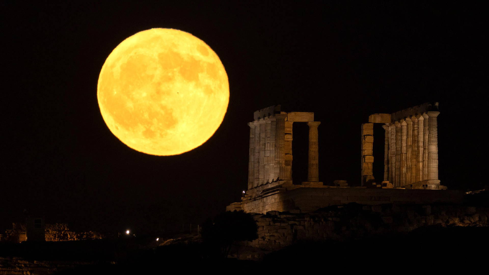 A yellow full moon is pictured looming large next to the columns of a ruined Greek temple, which is illuminated softly by artificial light.