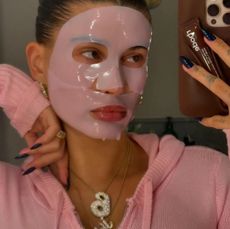 Hailey Bieber taking a selfie wearing a pink face mask and sweater with two diamond necklaces