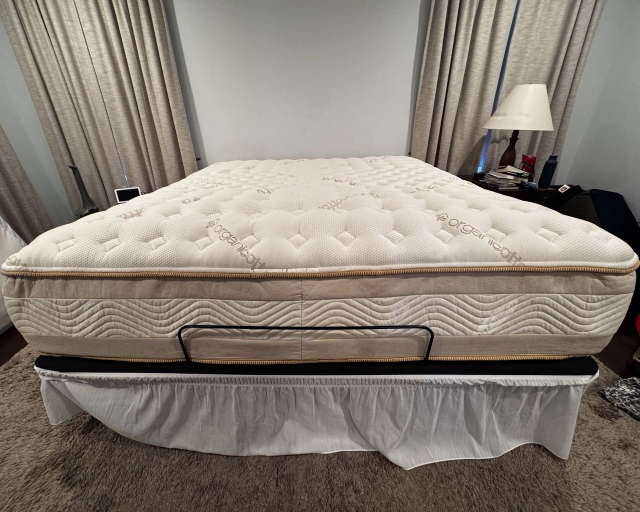 A bare, tan Saatva RX mattress on a platform with a white bed skirt. The bed is in a room that is painted blue with tan curtains.