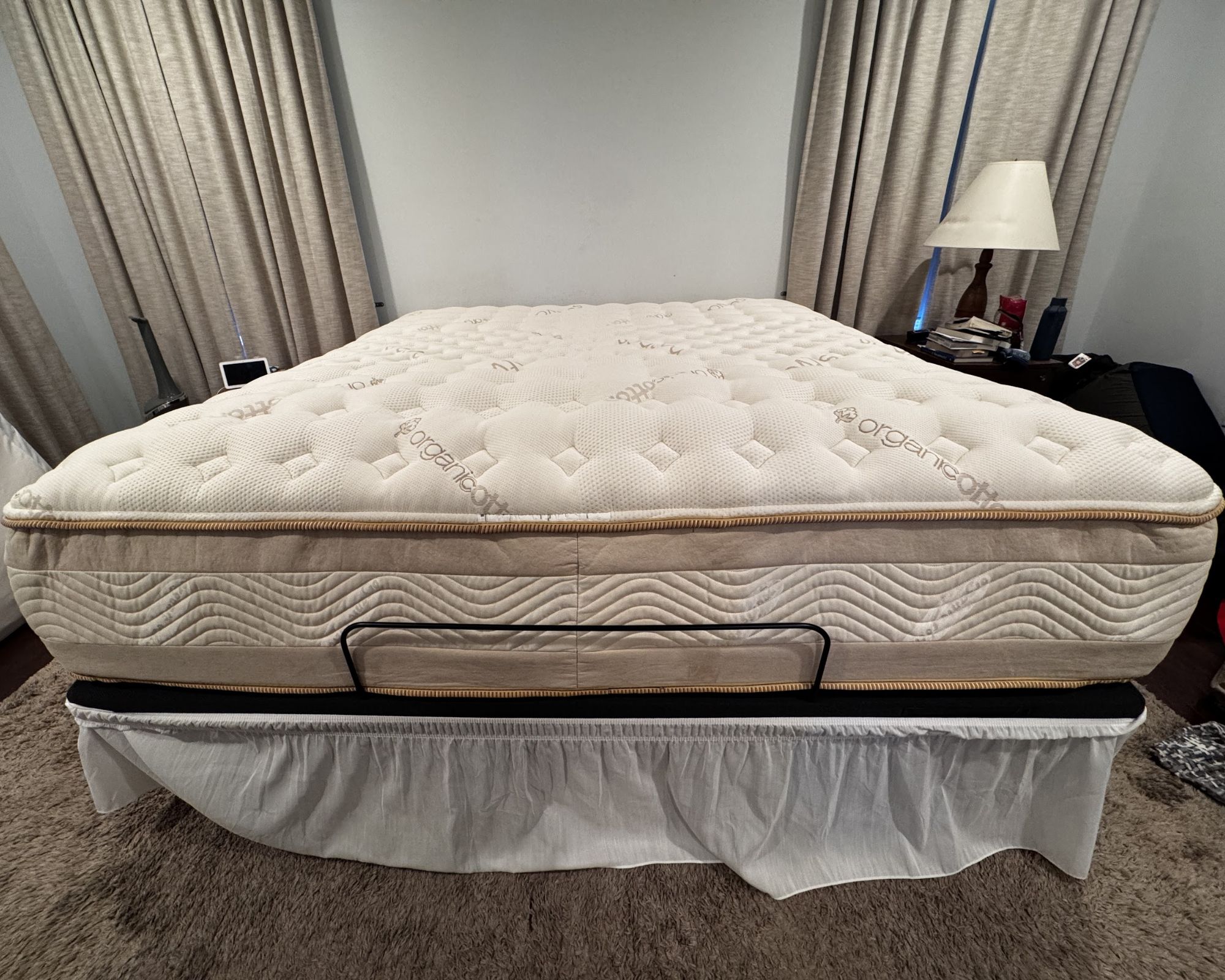 A bare, tan Saatva RX mattress on a platform with a white bed skirt. The bed is in a room that is painted blue with tan curtains.
