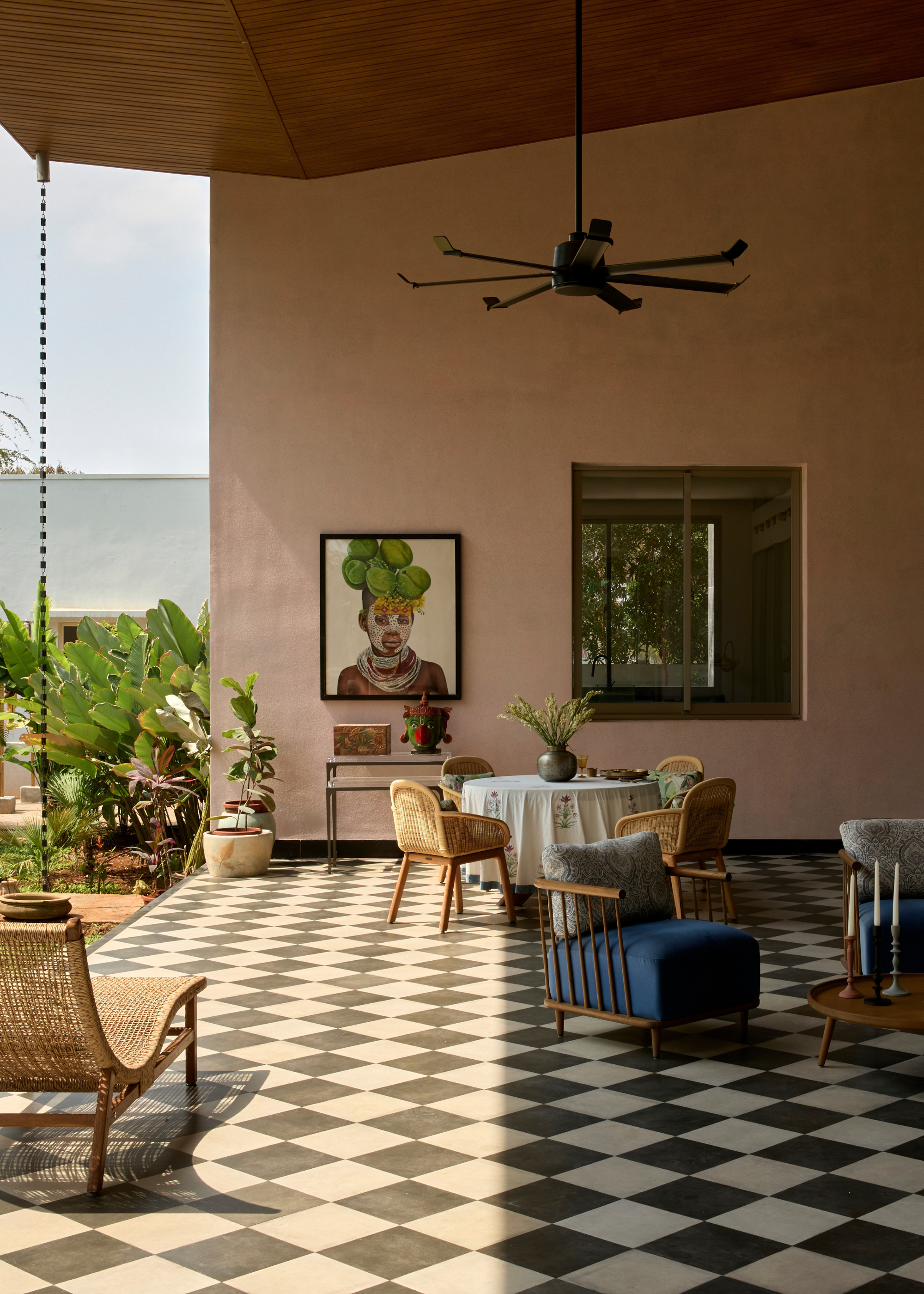 An outdoor space with checkerboard flooring, a rattan daybed, a group of wood chairs with pillows and a dining space with woven chairs below a fan