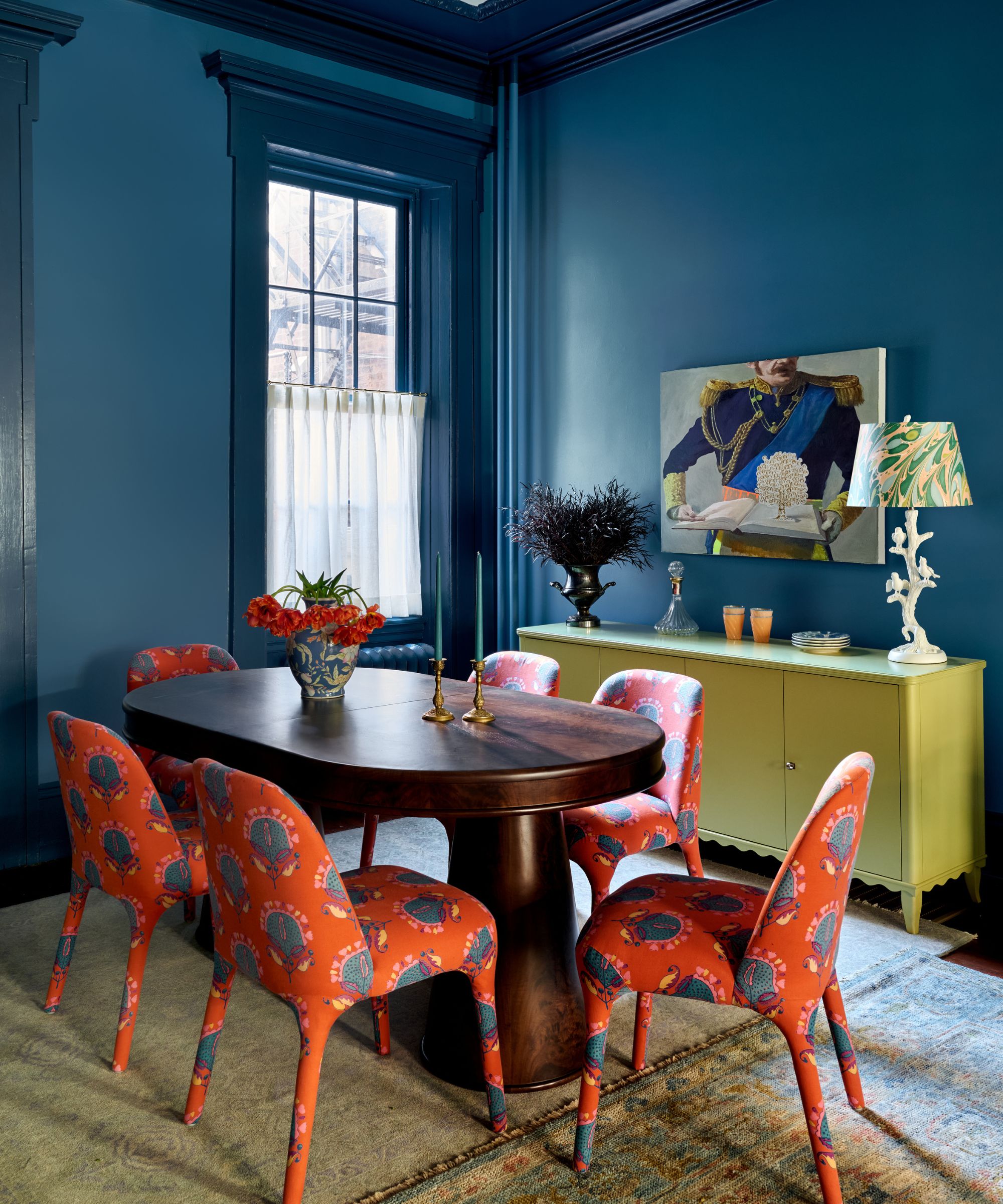 Dining room with deep teal walls, an oval dark wood table, six bright coral paisley chairs, and an olive-green credenza.