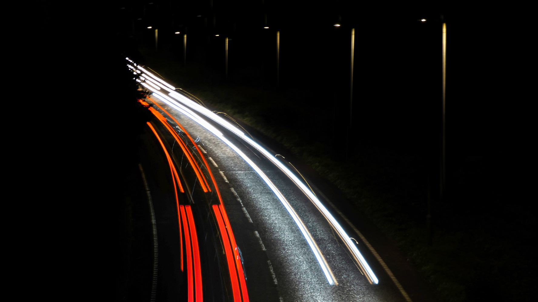 Light trails of cars on a dark road, illuminated by street lamps, creating a vibrant, dynamic pattern against the night sky