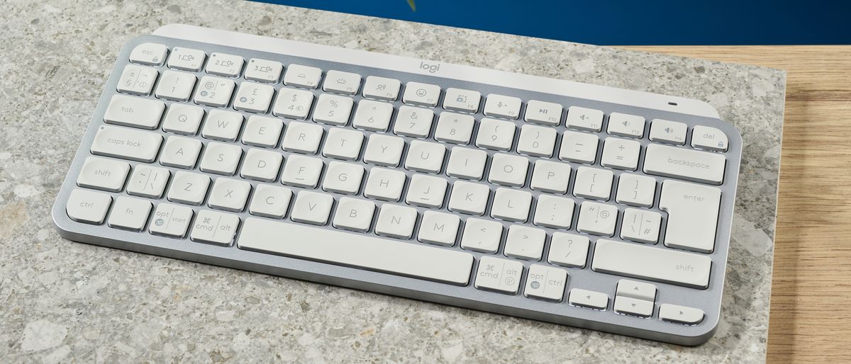 A photograph of the Logitech MX Keys Mini in light gray, positioned on a stone slab with a blue wall in the background.