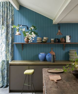 A blue panelled kitchen with patterned curtain, open shelving, green and wooden countertops and green stool