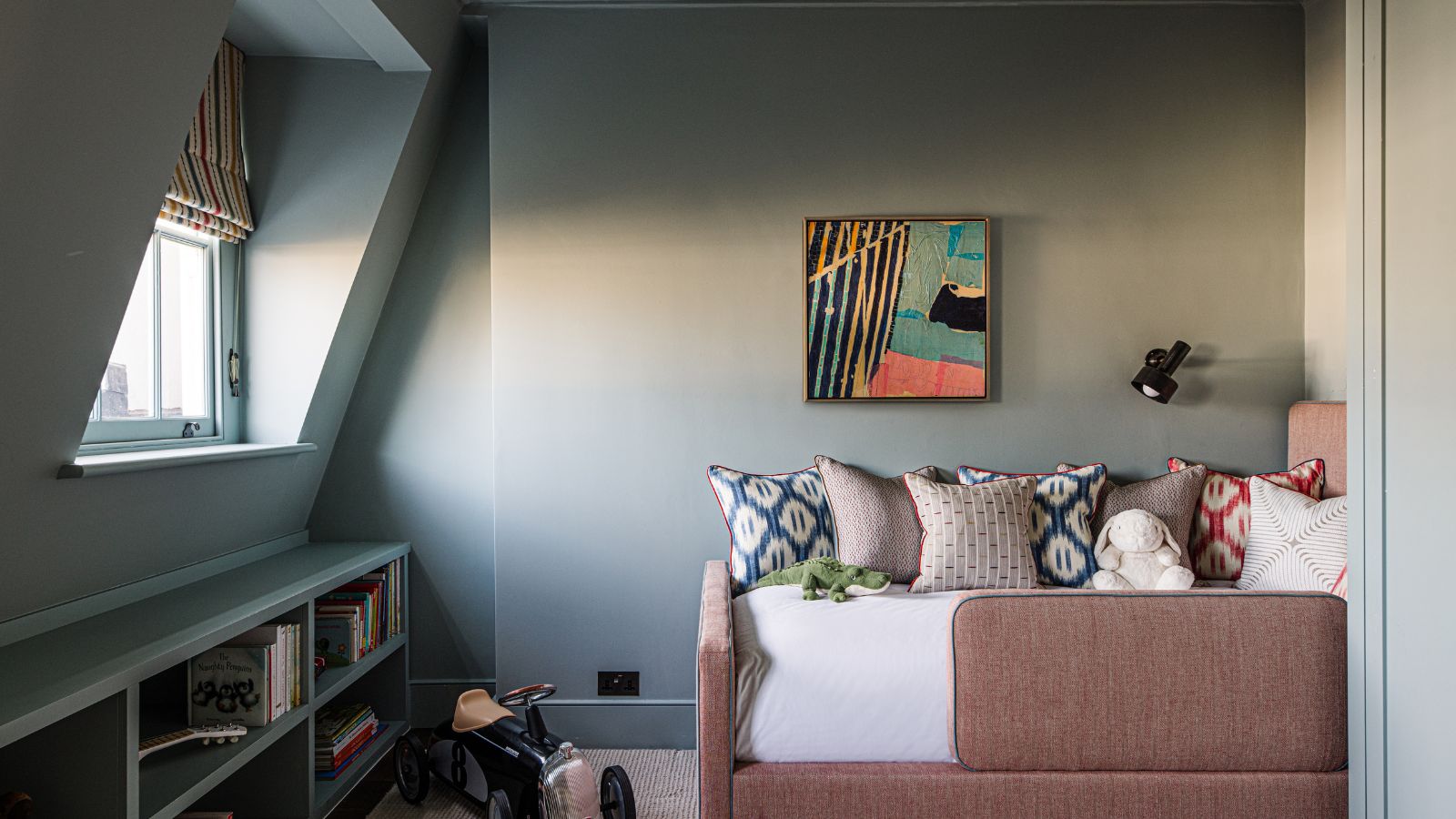 Gray Blue attic bedroom for kid with striped blinds, built-in bookshelf, artwork on the wall, and pink bed with lots of cushions