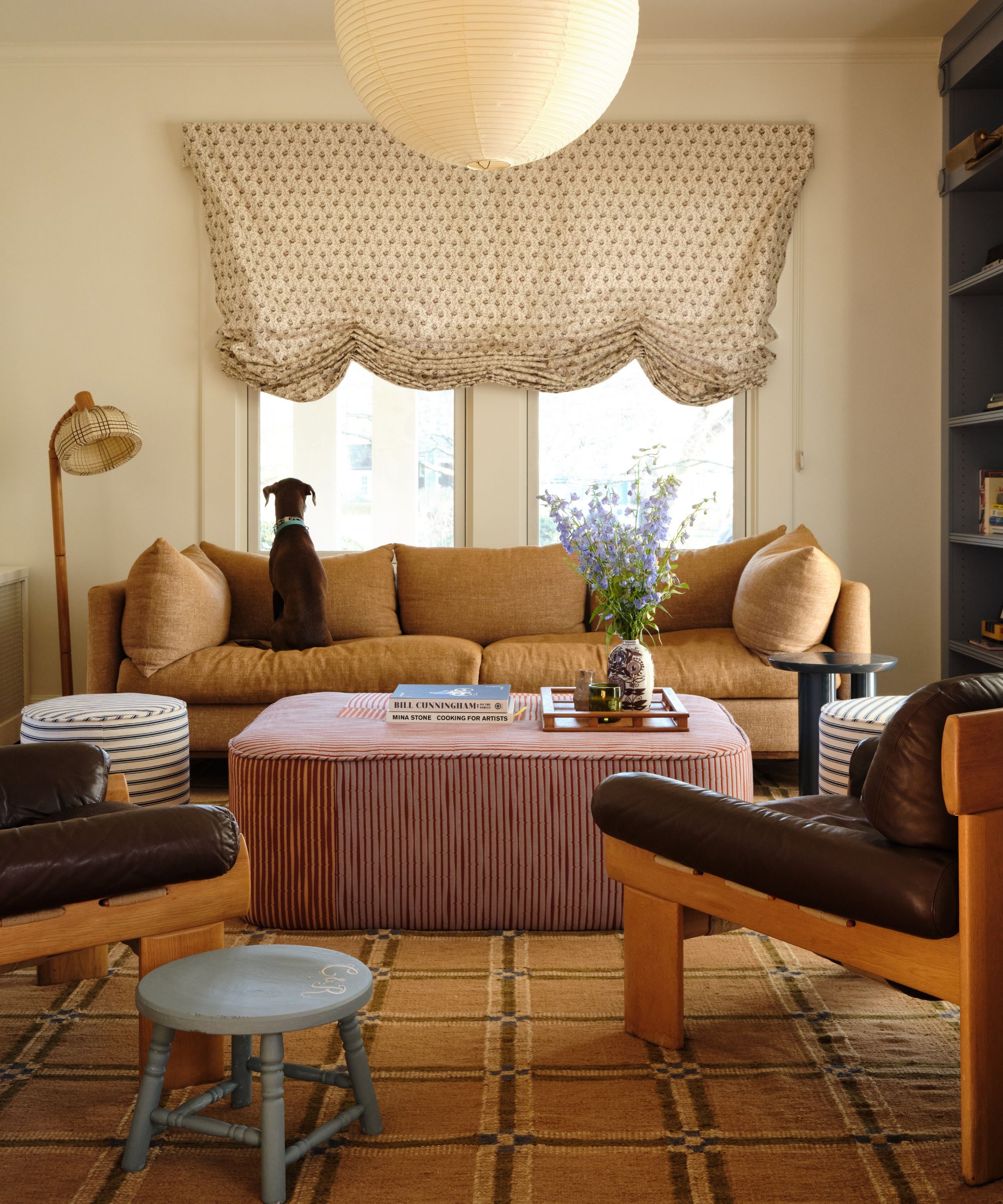 Living room with balloon blind, beige plaid carpet, striped ottoman, midcentury leather lounge chairs, pale blue wooden stool and beige linen couch with dog perched on top