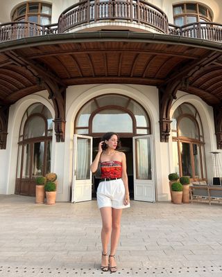 Melodie wears long pleated shorts and silky red top with black strappy heels.