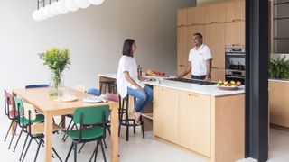 A couple in a large open plan minimalist kitchen with wood cabinets, long dining table, and large island breakfast bar