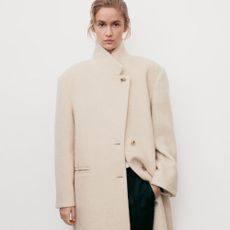 Woman wears cream coat with high collar, black trousers