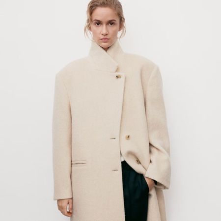 Woman wears cream coat with high collar, black trousers