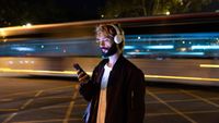 A young, bearded man stands looking at his phone at night while wearing over-ear headphones with a blurred train going past in the background. 