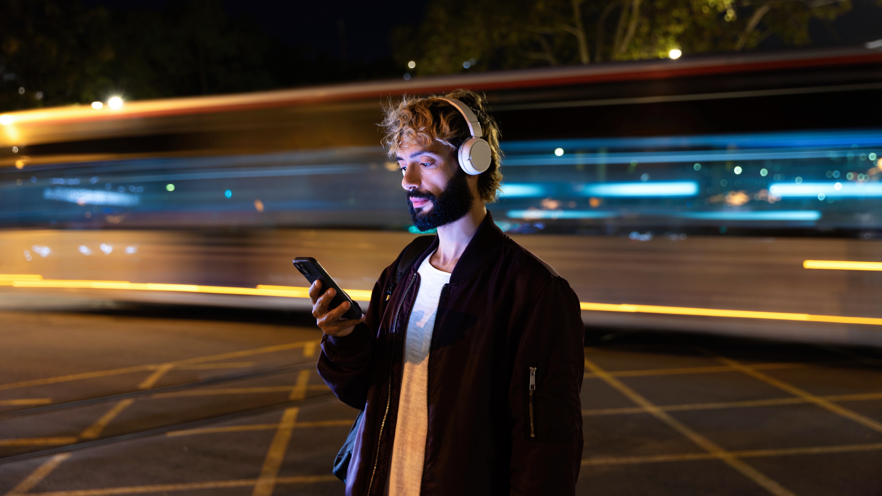 A young, bearded man stands looking at his phone at night while wearing over-ear headphones with a blurred train going past in the background. 