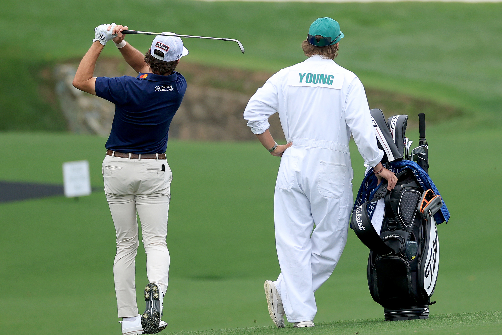 Cameron Young hitting an iron shot at Augusta National Golf Club during a practice round for the Masters, next to his caddie bearing his name on the famous white boiler suit