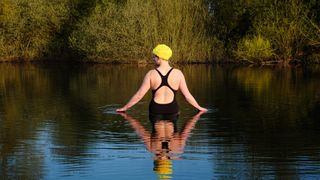 UK, Essex, female wild swimmer standing in a lake touching the water at sunrise