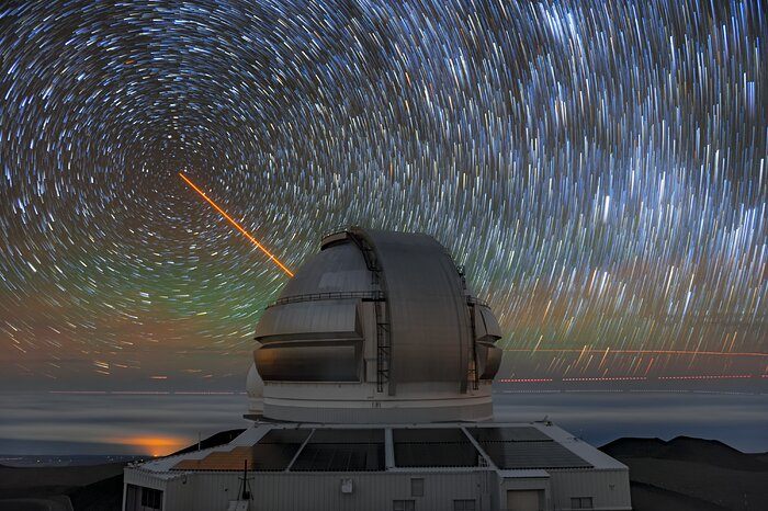 Un láser naranja dispara desde el lado opuesto de una estructura circular de observatorio, apuntando hacia el norte mientras las estrellas orbitan alrededor de su eje apuntando a la Estrella Polar, dispersada por una larga exposición de la cámara.