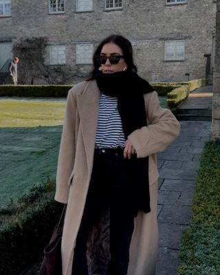 Influencer wears camel coat, black scarf, striped shirt, and black jeans.