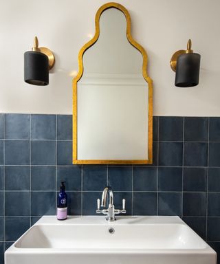 bathroom with blue square tiled lower half, white sink, gold shaped mirror, black wall lights