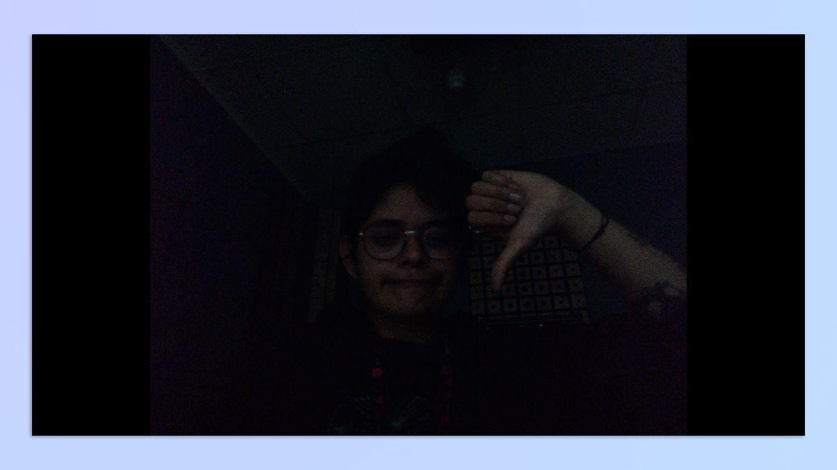 A woman with short hair sitting in a dark room, holding up her hand in the shape of a thumbs-down