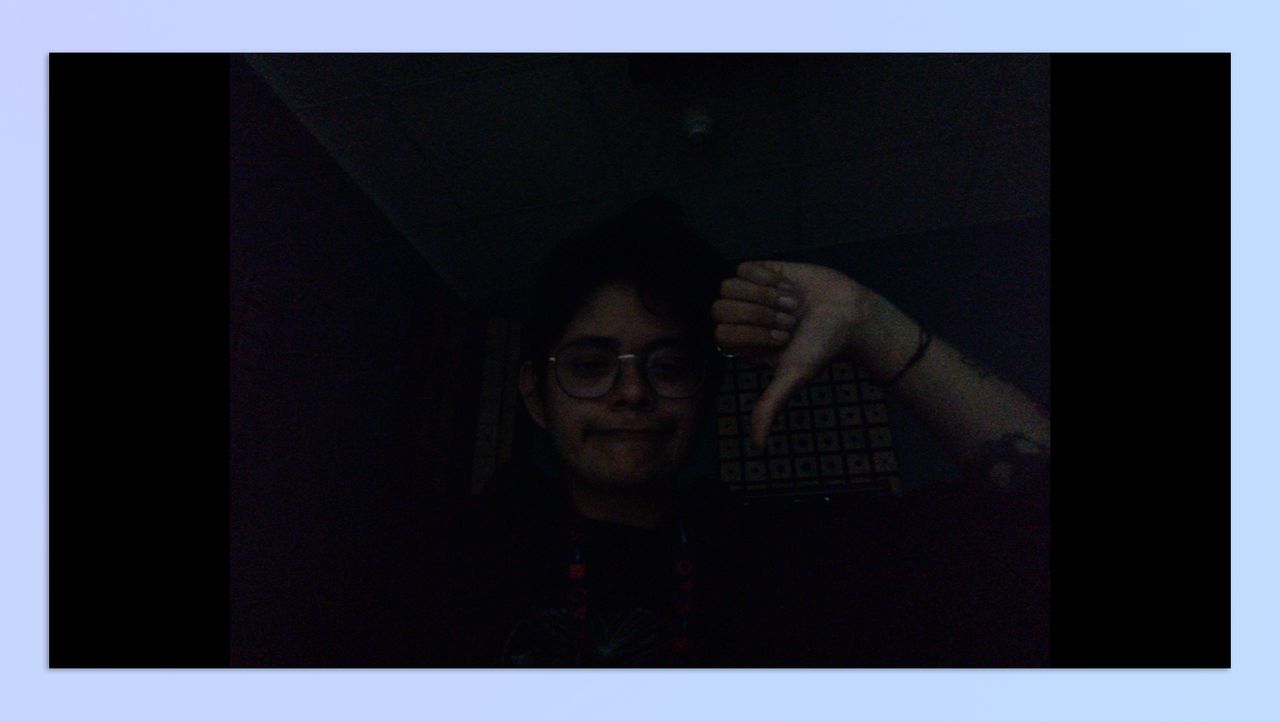 A woman with short hair sitting in a dark room, holding up her hand in the shape of a thumbs-down