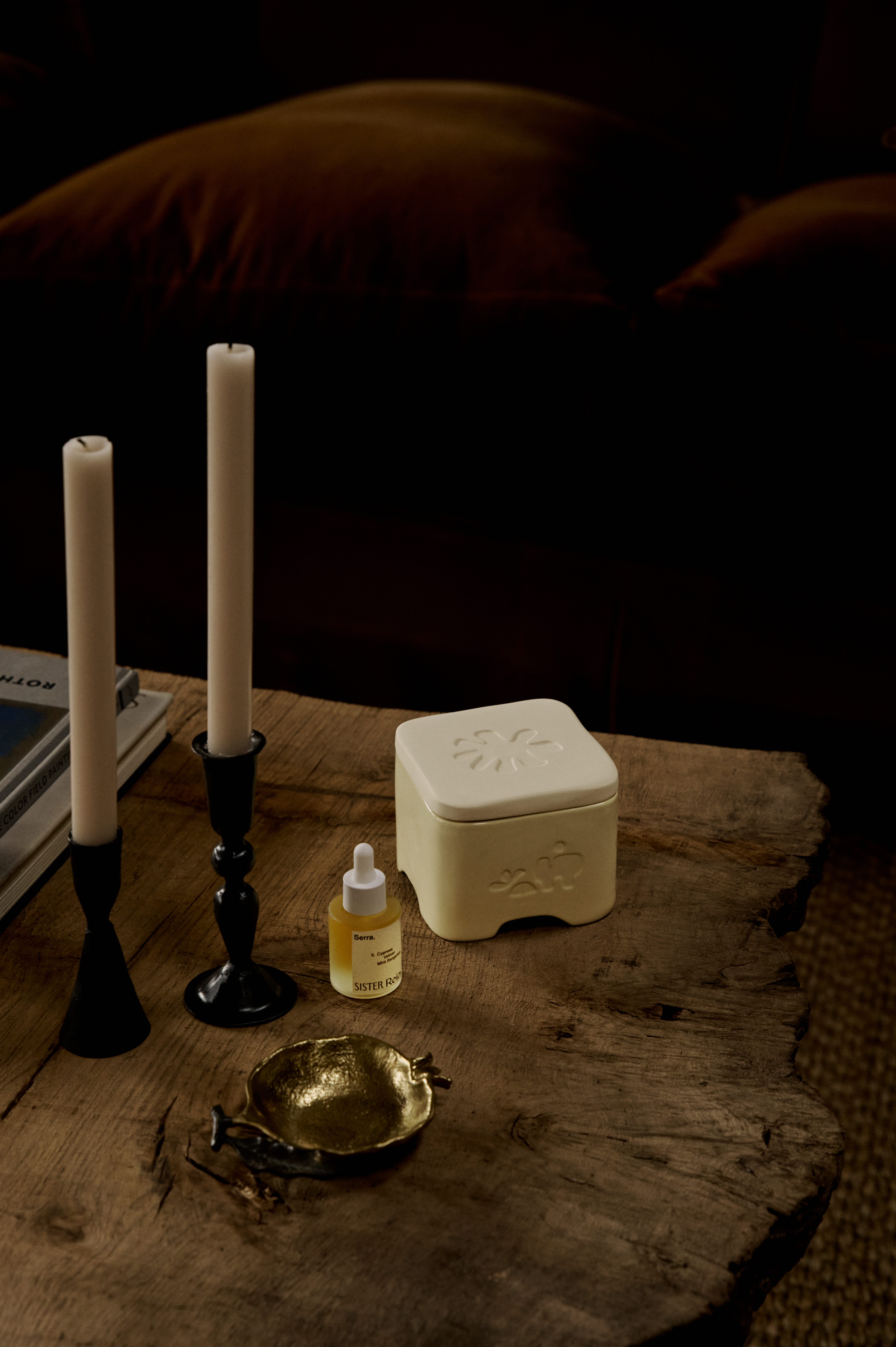 A room lit by a sunset boasts a home fragrance diffuser in the form of a pale yellow box, with a bottle of perfume, a vase of flowers, books, and other collectibles like candles and home accessories.