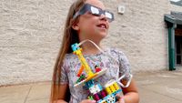 a young girl wears eclipse glasses while looking up and holding a lego creation