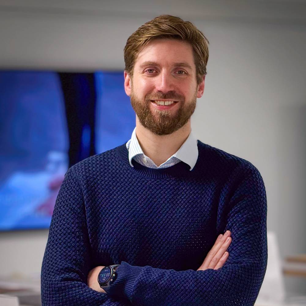 male with short hair and beard wearing navy jumper and white shirt, stood wth arms folded in office setting