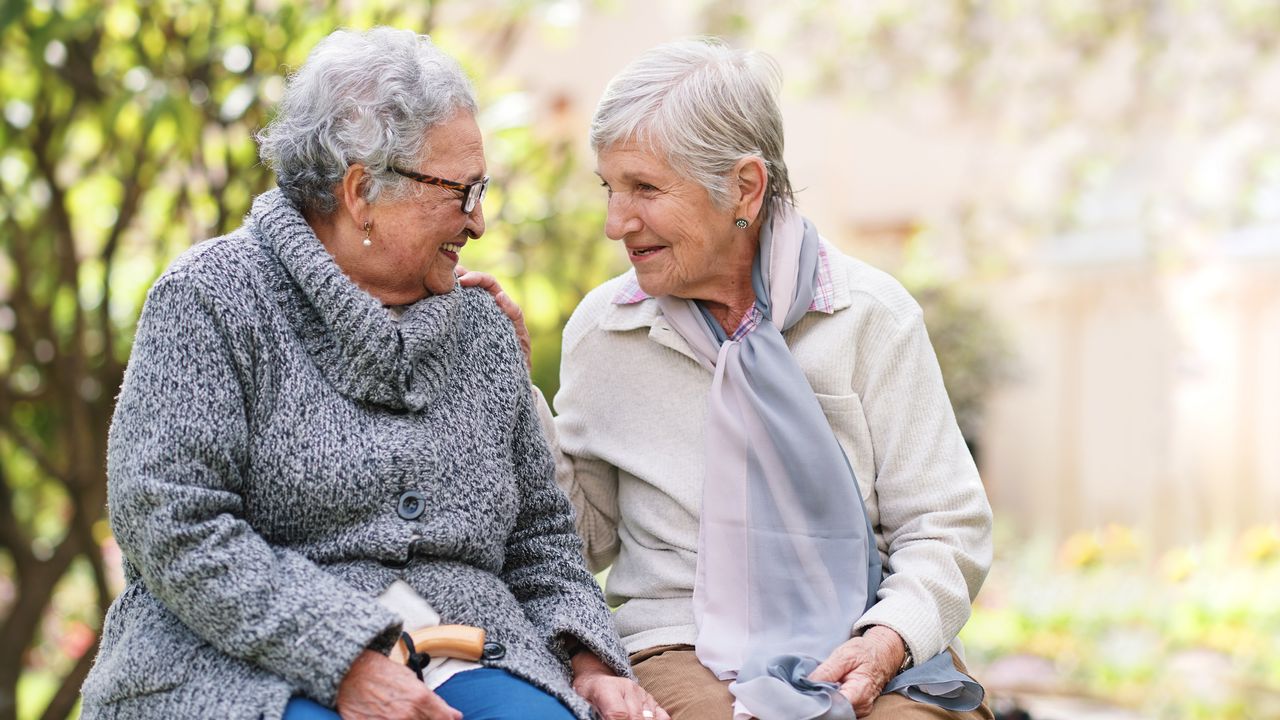 Two older women sit on a bench outside and talk and smile.