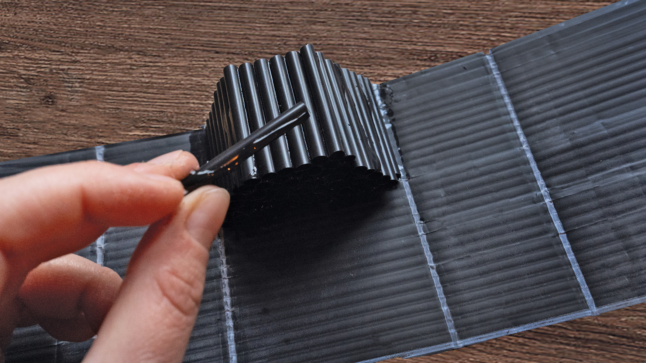 A hand holding a black stick applies glue to a textured, ridged black structure on a textured dark surface