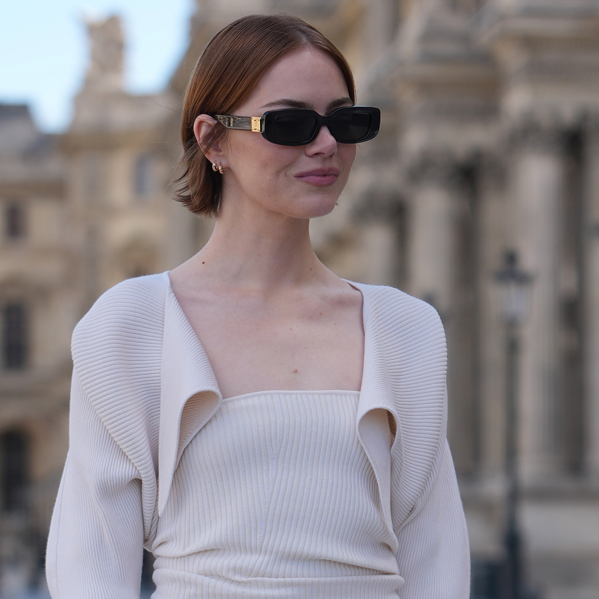 PARIS, FRANCE - SEPTEMBER 30: Emma Stone wears auburn chin-length straight hair with a center part, black rectangular sunglasses with gold metal temple detailing, gold hoop earrings and a gold ring, a black leather satchel bag with top handles and silver hardware carried handheld, a cream ribbed knit strapless midi dress with a ruched bodice and fitted pencil skirt layered with a matching cream ribbed knit bolero-style cropped long-sleeve cardigan with an open draped front, black leather pointed-toe pumps shoes with stiletto heels, outside Louis Vuitton, during Paris Fashion Week Womenswear Spring/Summer 2026, on September 30, 2025 in Paris, France 