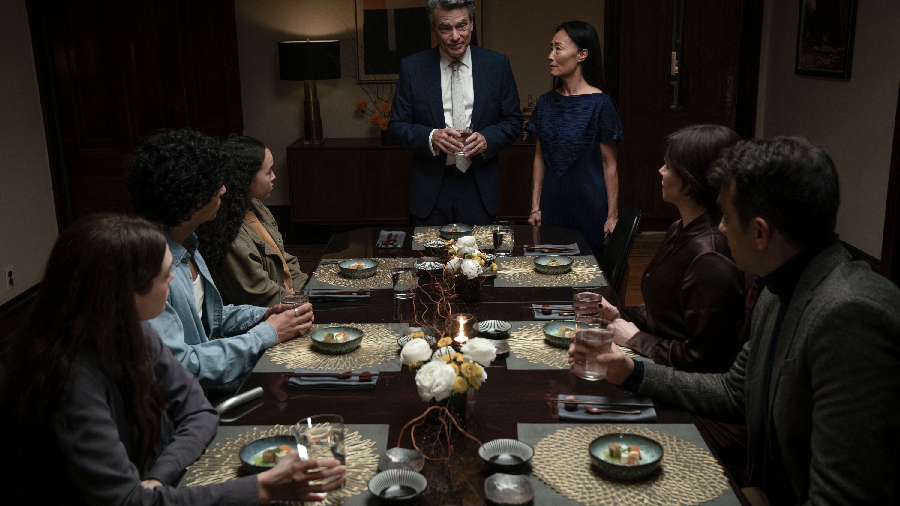 a family sits around a table looking at a man (peter gallagher) and woman standing at the head, in a still from 'humane'