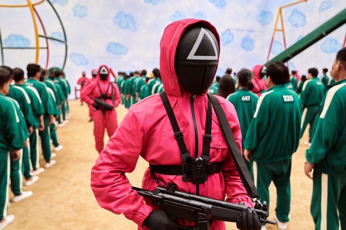 Masked men carrying guns between lines of contestants in jumpsuits.