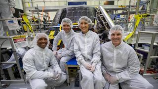 four people in white cleansuits and hair nets smile in front of a cone-shaped spacecraft in a laboratory