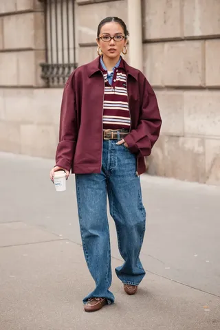 A woman wears a burgundy coat, a striped button down shirt, brown belt, baggy jeans, and brown boat shoes.