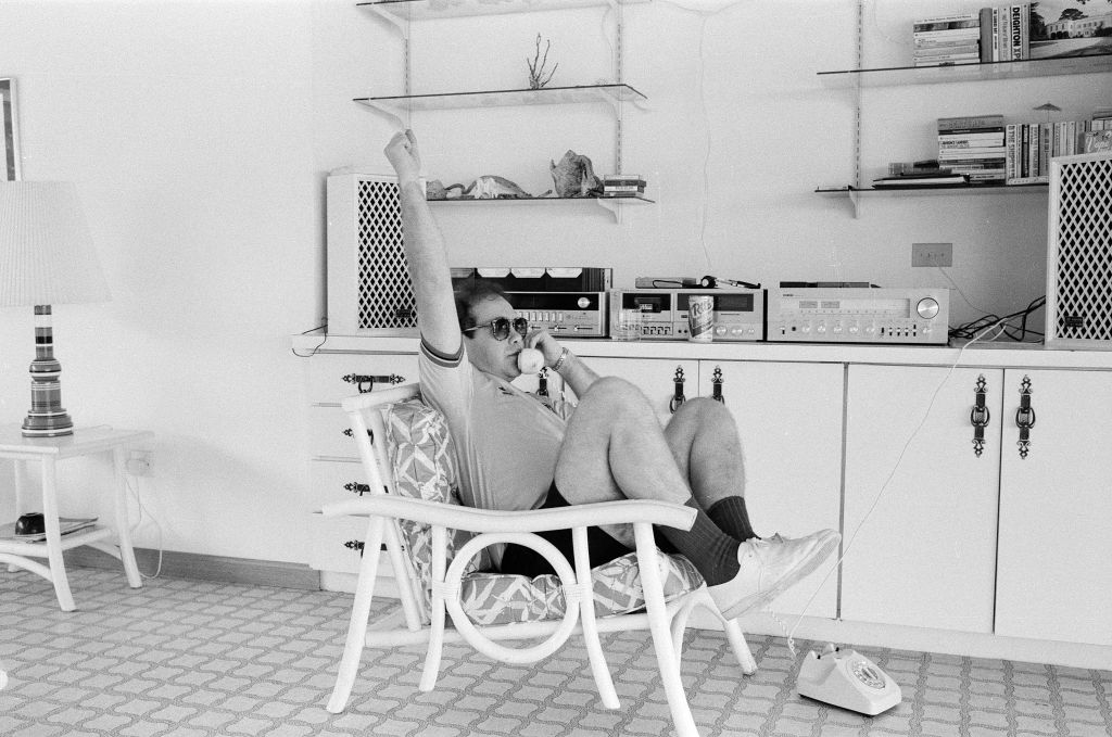 Elton John on the Caribbean island of Montserrat to record his next album (Too Low for Zero), pictured taking taking a break to listen in to his beloved football team Watford FC play, arm raised as he celebrates a goal, October 1982. (Photo by Carl Bruin/Mirrorpix via Getty Images)