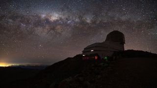 the silhouette of an observatory sits atop a dark hill beneath the milky way stretched across a starry sky.