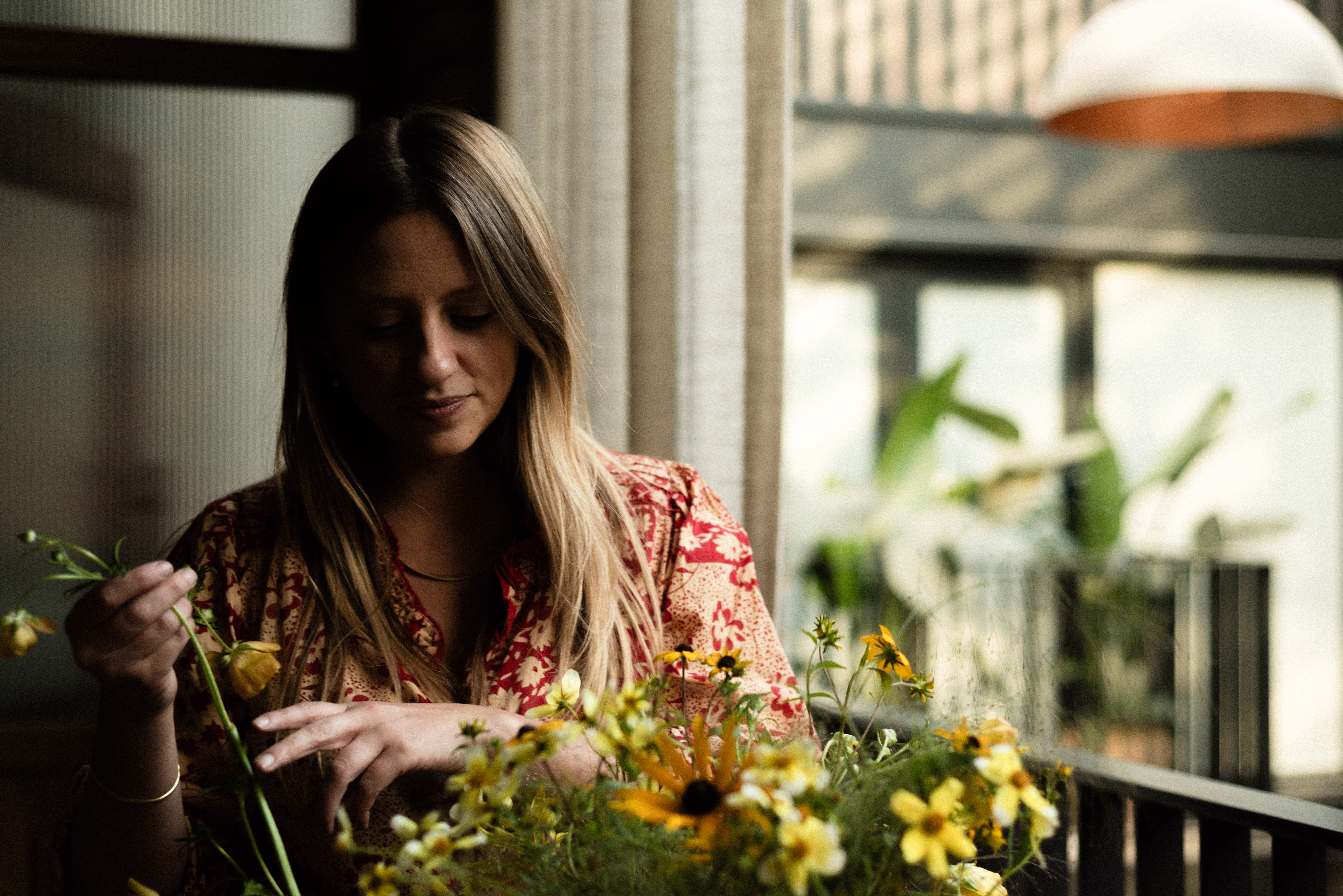 A young woman in a cream and red floral blouse handles yellow flowers in a restaurant whose interior gives onto a sun-lit piazza.