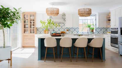 A white kitchen with blue island, taupe bar chairs, white and blue patterned tiles and wooden glass-fronted cabinet 