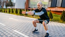 A man in sportswear performs a banded lateral walk outside. He has a short looped resistance band wrapped around his thighs and he is squatting down, with knees bent and thighs parallel to the floor. He holds his arms straight out in front of him. Behind him we see a building with large windows and a line of young fir trees.