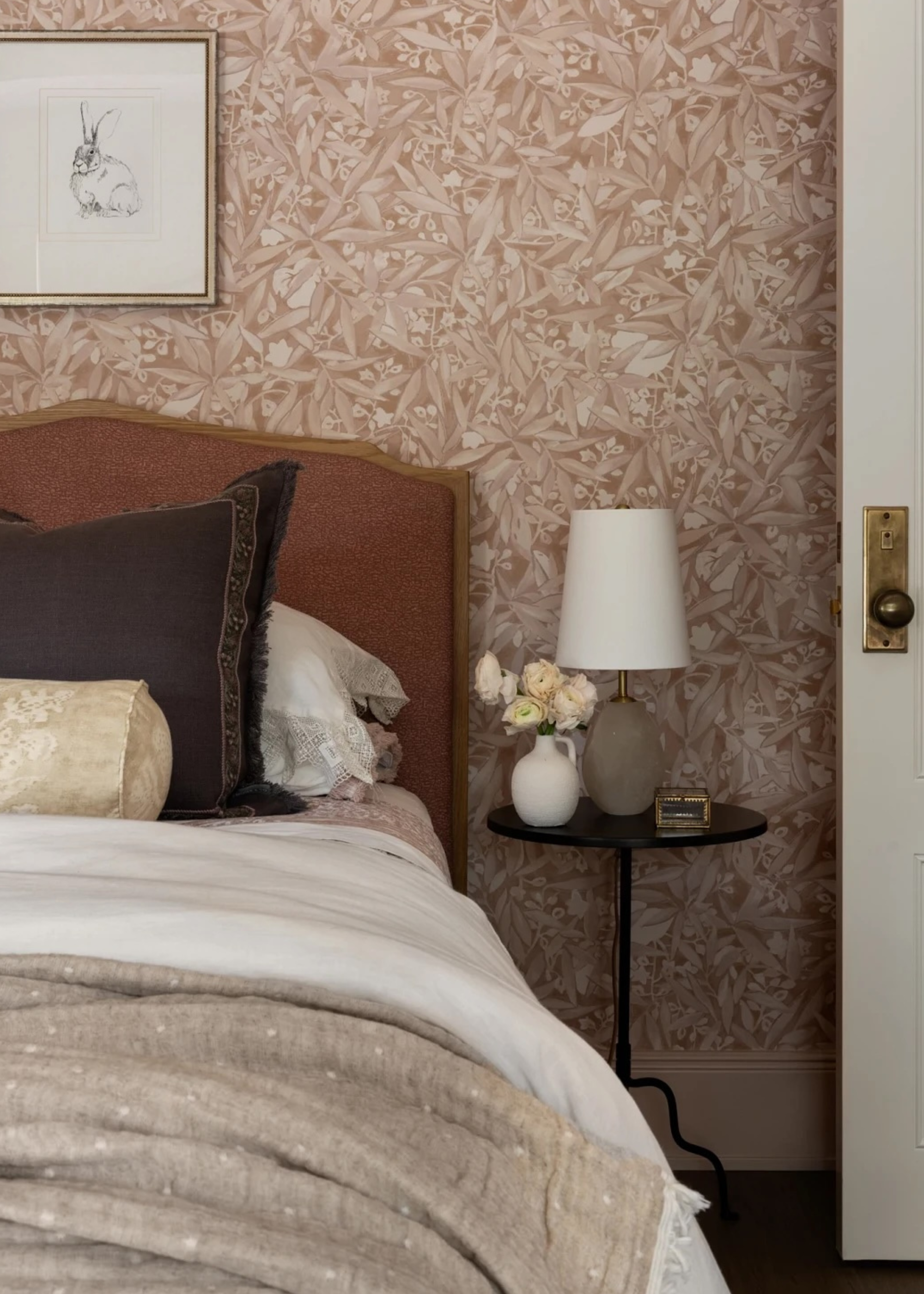 A bedroom with a sandy wallpaper, a framed bunny sketch, a mustard headboard, a bed with a stack of pillows, a side table with a lamp and a vase of flowers