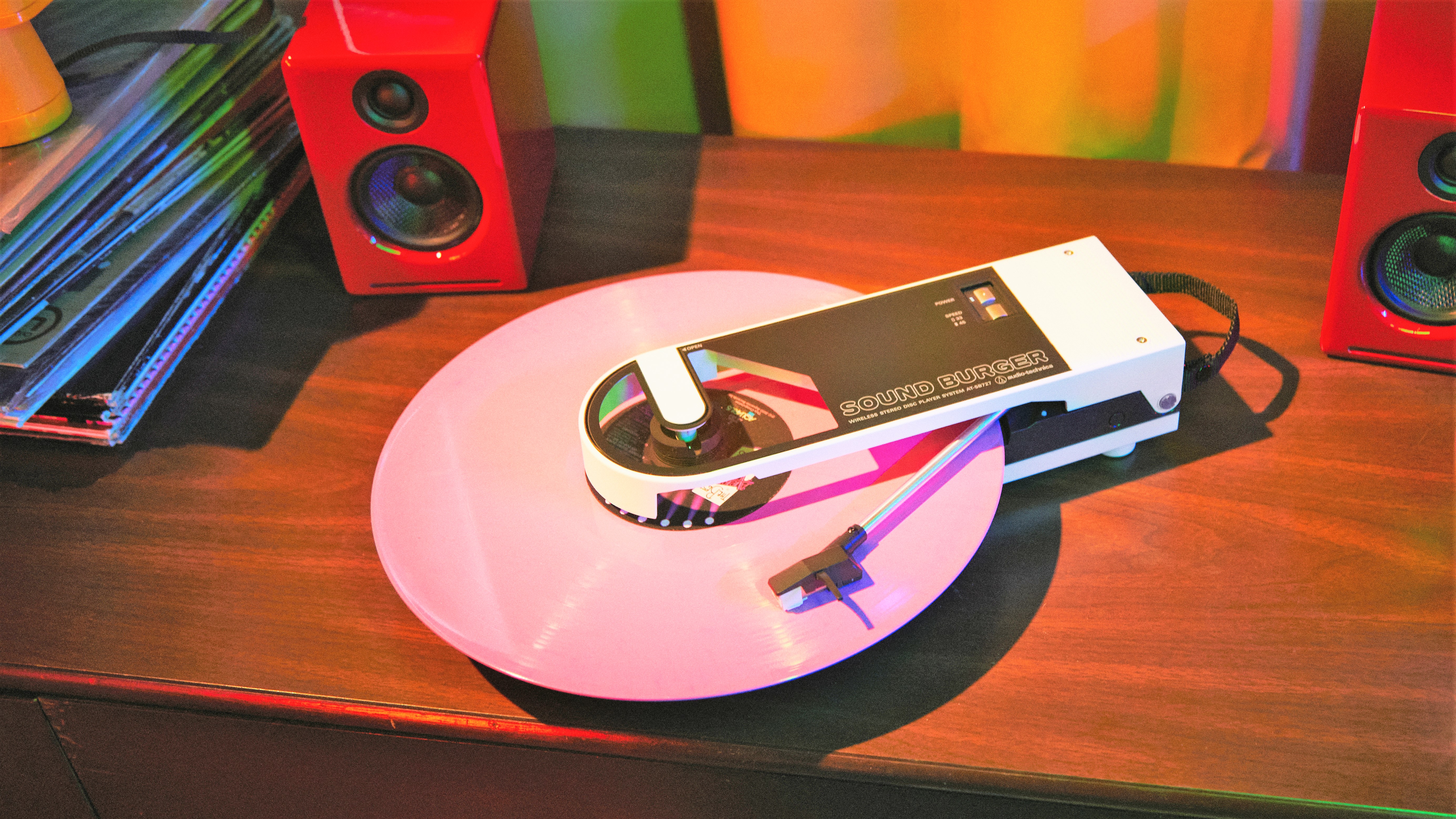 Audio-Technica Sound Burger spielt eine rosafarbene Schallplatte auf einem Schreibtisch mit LPs und Lautsprechern im Hintergrund