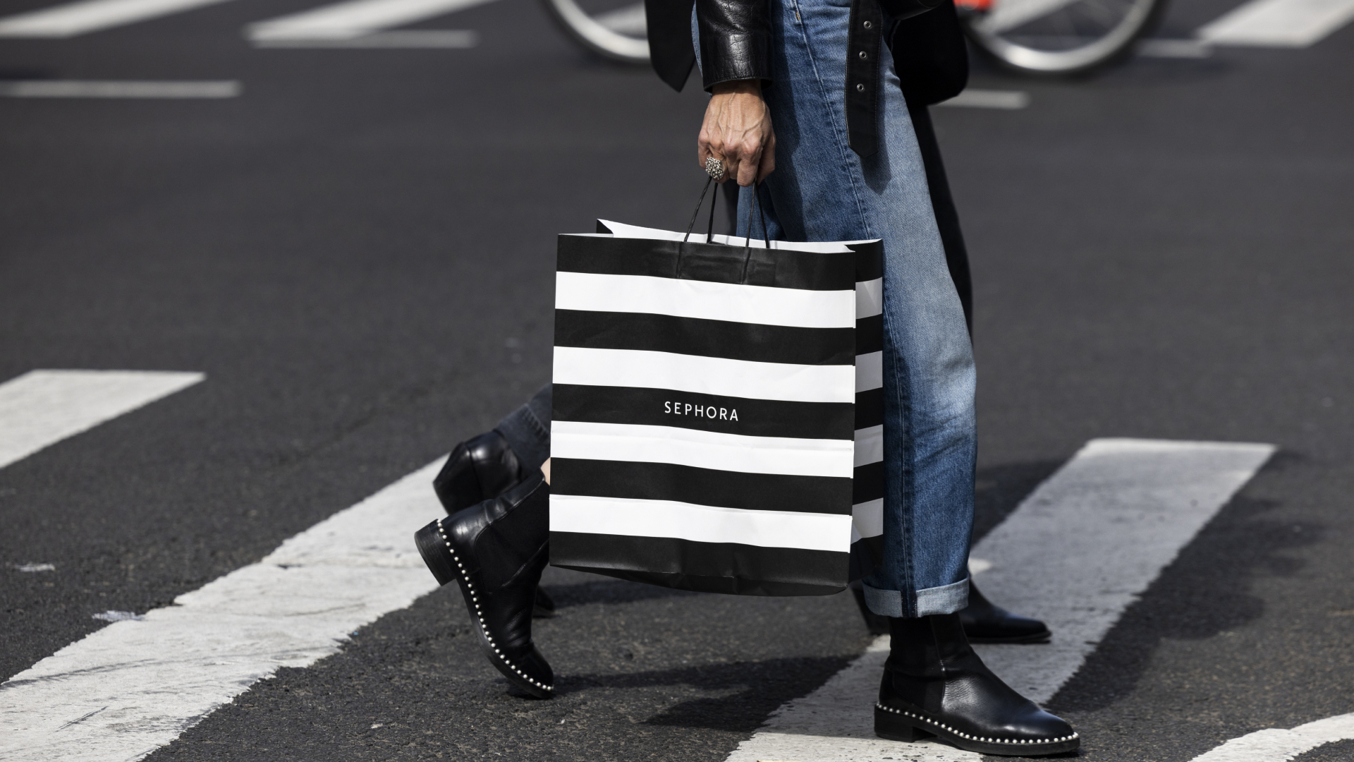 A person photographed from the waist down wearing blue jeans and black boots carrying a black and white striped Sephora shopping bag