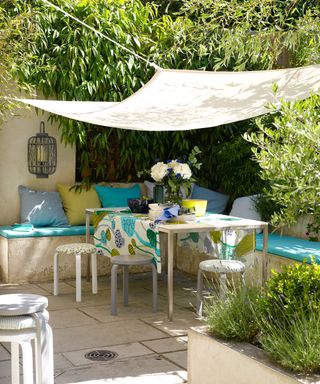 The corner of a patio draped with a sail shade and built-in bench seating, table and stools decorating with white, green and blue soft furnishings.
