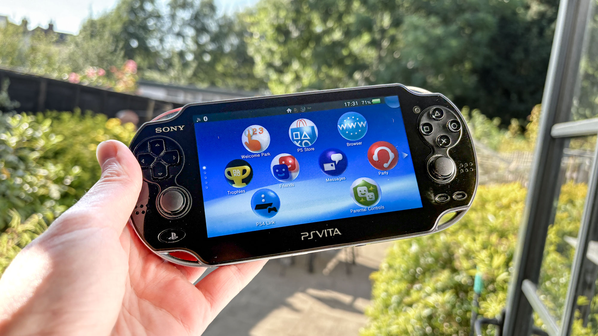 PS Vita in hand with outside garden in background with homescreen menu on display