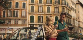 Miranda (Elen Rhys) and Max (Julian Looman) lean back against an open-topped car in the sunshine in the streets of Palma. Both of them are eating ice lollies, and Max is holding a tablet computer.