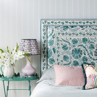 blue and white bedroom with upholstered headboard, teal metal side table, vase of flowers, pink lamp with patterned shade, pale blue and white wallpaper, pale blue bedding, pink and teal cushions
