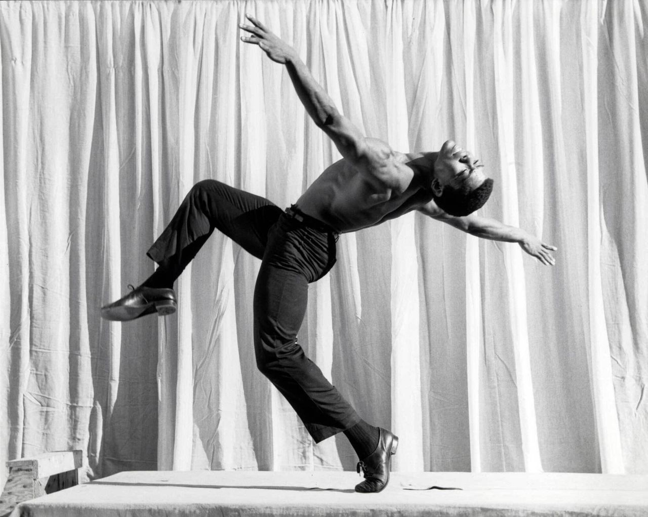 A dancing man is captured while performing on a stage in front of a white curtain.