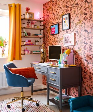 A colorful home office with yellow curtains, wooden wall shelf with decor, pink patterned wallpaper with wall art, a navy blue office chair, a dark gray desk with a laptop and papers and books on it